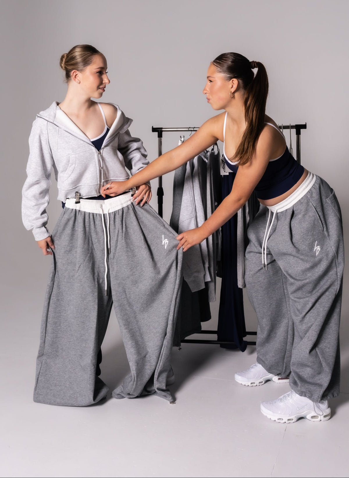 Two women modeling matching gray wide-leg sweatpants in a studio setting, with one holding out the fabric to show the fit and the other wearing the pants with a white sports bra and white sneakers.