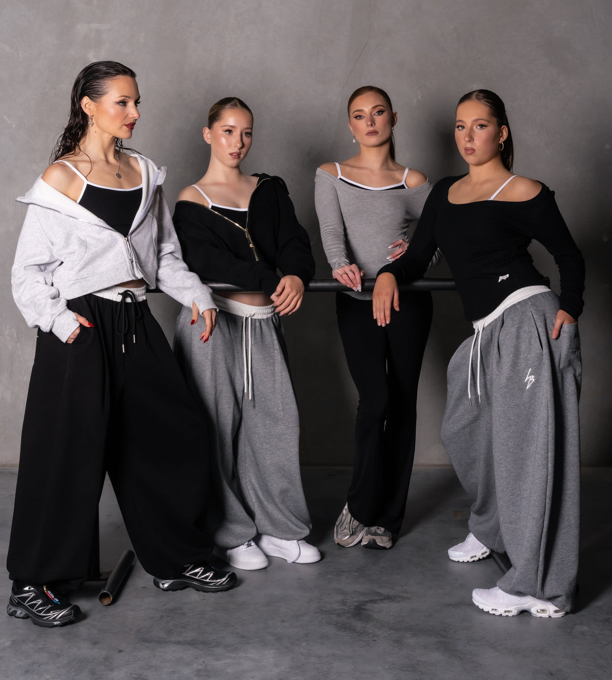 Four women modeling different streetwear outfits in a studio setting with a gray background. The outfits include black and white, black and gray, gray and black colorways, featuring off-the-shoulder tops, wide-leg sweatpants, and sneakers.