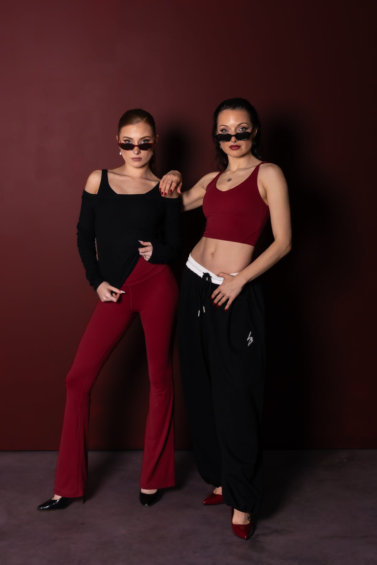 Two women posing in stylish outfits against a dark burgundy background. The woman on the left wears a black cold-shoulder top and red flare pants, while the woman on the right wears a matching red sports bra and black baggy sweatpants.